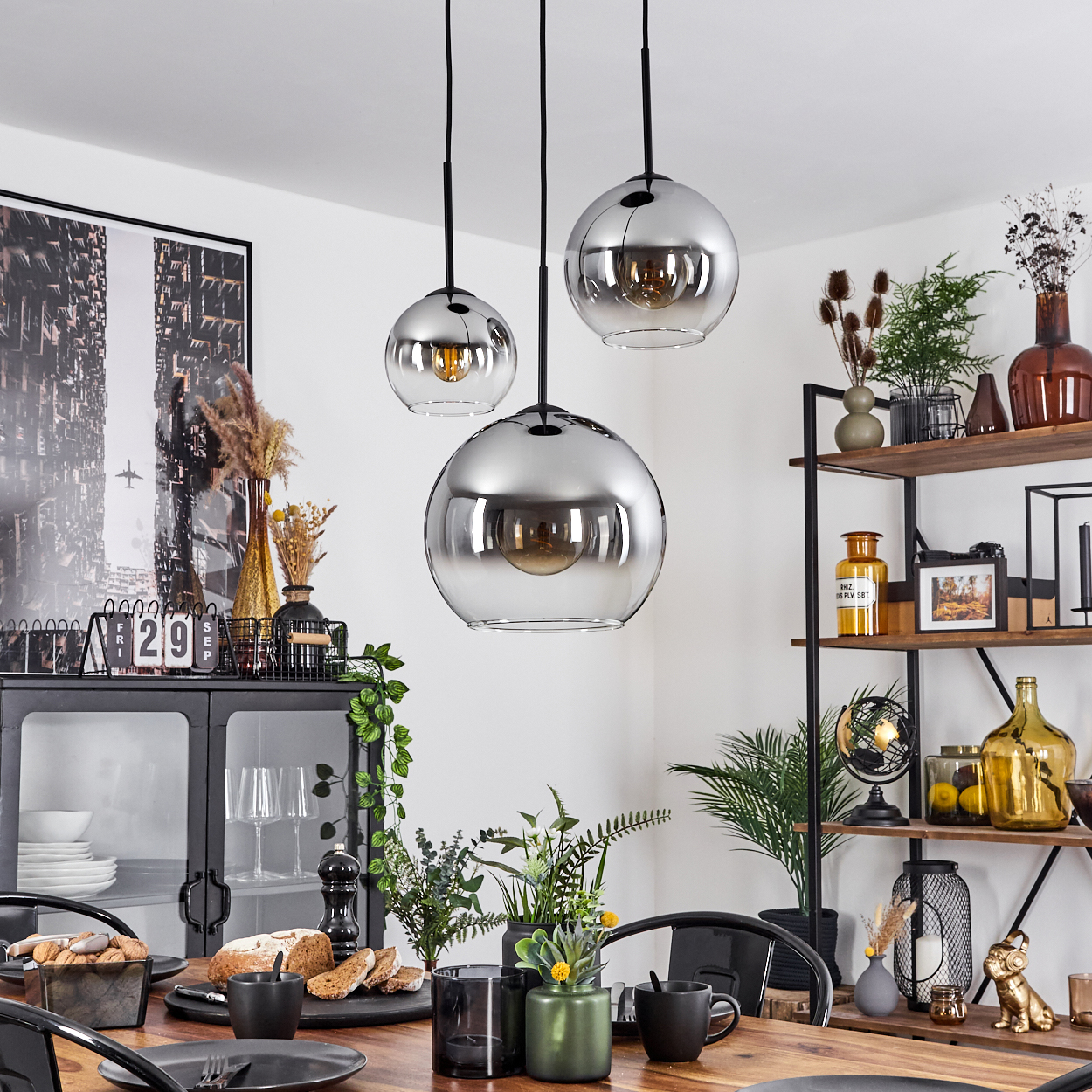Esszimmer-Szene. Drei Pendelleuchten über einem Holztisch. Dekoration umfasst Pflanzen, Kunst und einen schwarzen Schrank.