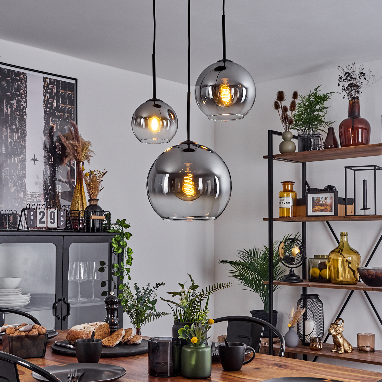 Esszimmer-Szene. Drei Pendelleuchten, Holztisch, schwarzer Schrank, Pflanzen und Kunst. Die Lampen sind Chromkugeln.