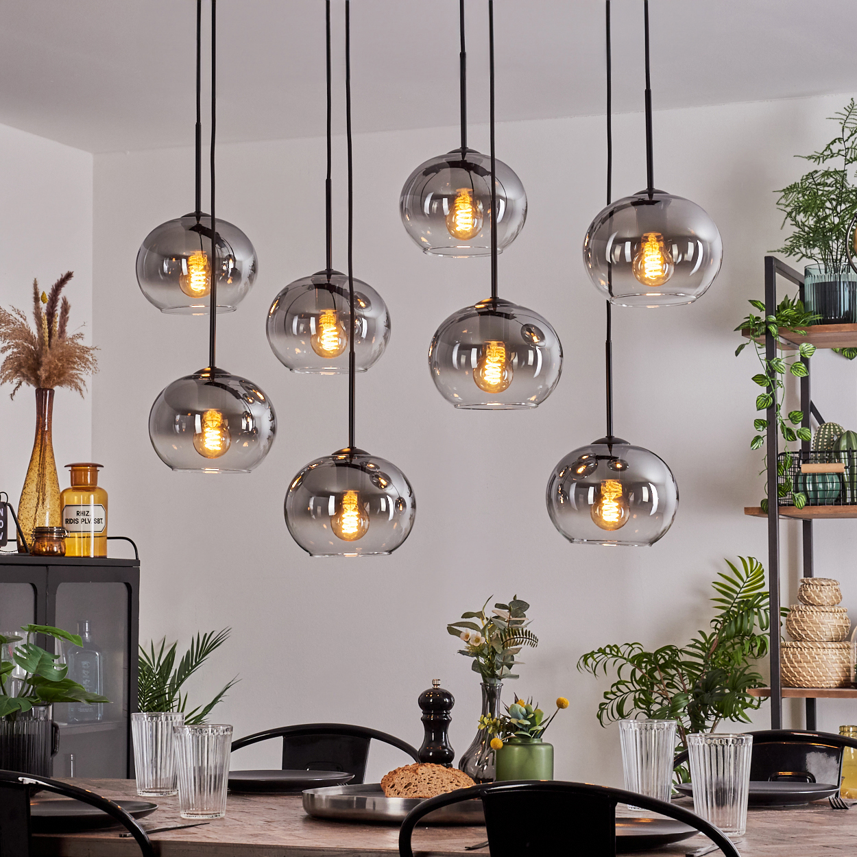 Esszimmer mit acht rauchgrauen Glaskugel-Pendelleuchten über einem Holztisch. Pflanzen und Glaswaren sind sichtbar.