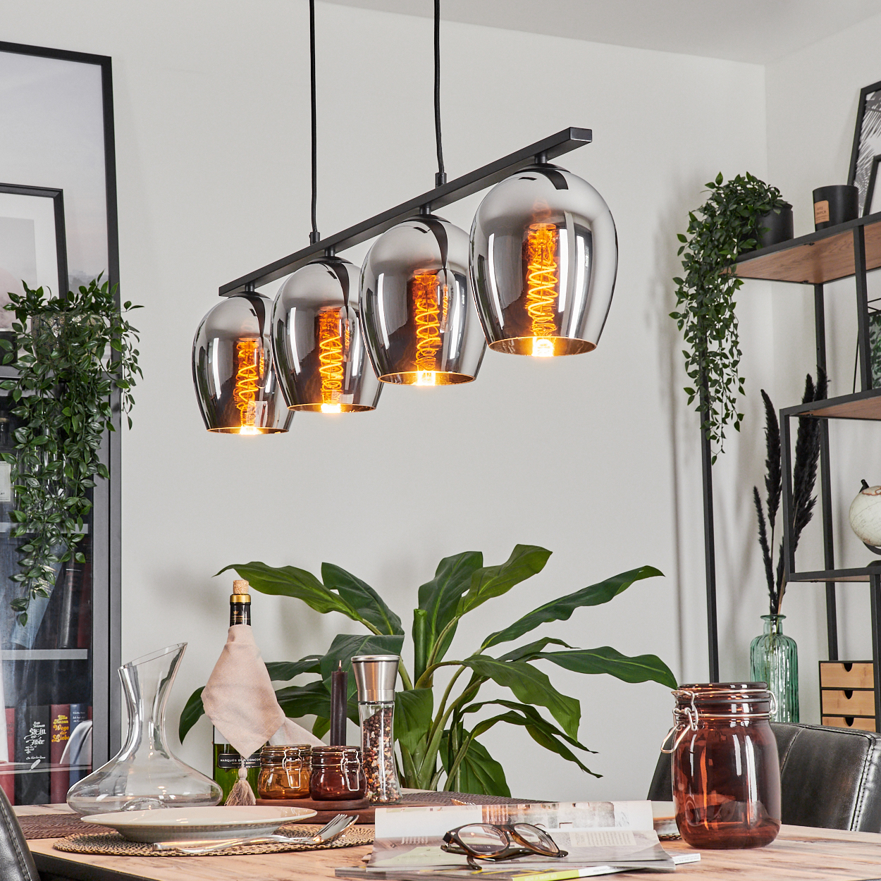Esszimmer mit Holztisch, vier Pendelleuchten mit orangefarbenen Glühbirnen und verschiedenen Dekorationen. Eine große Pflanze ist vorhanden.