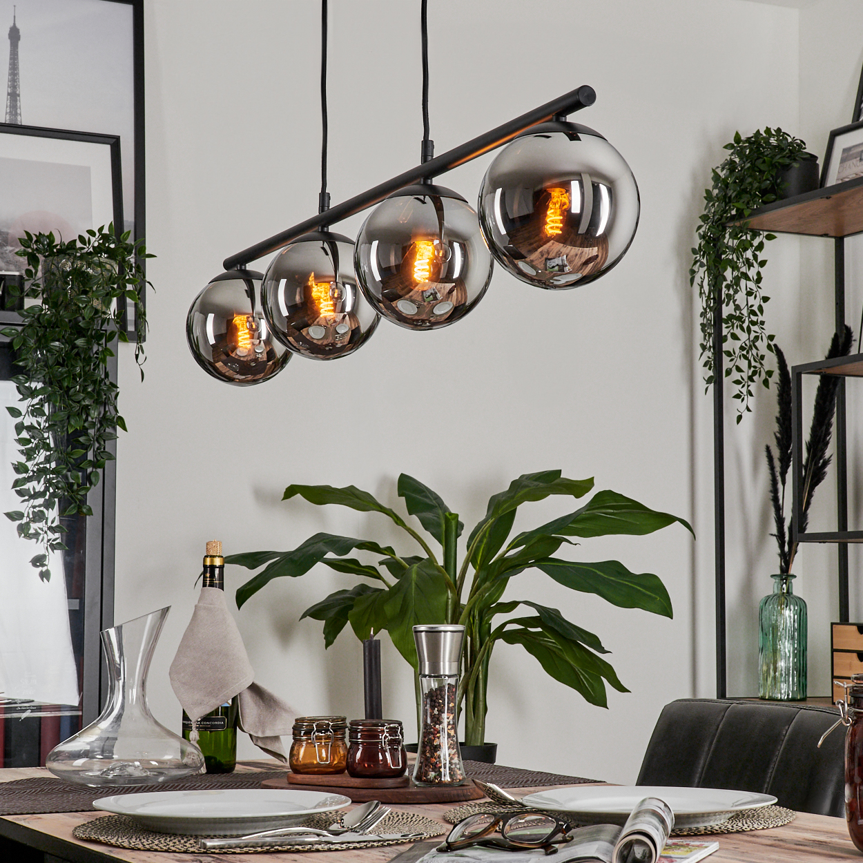 Esszimmer mit Holztisch, vier Pendelleuchten mit orangefarbenen Glühbirnen und verschiedenen Dekorationen. Die Lampen sind an.