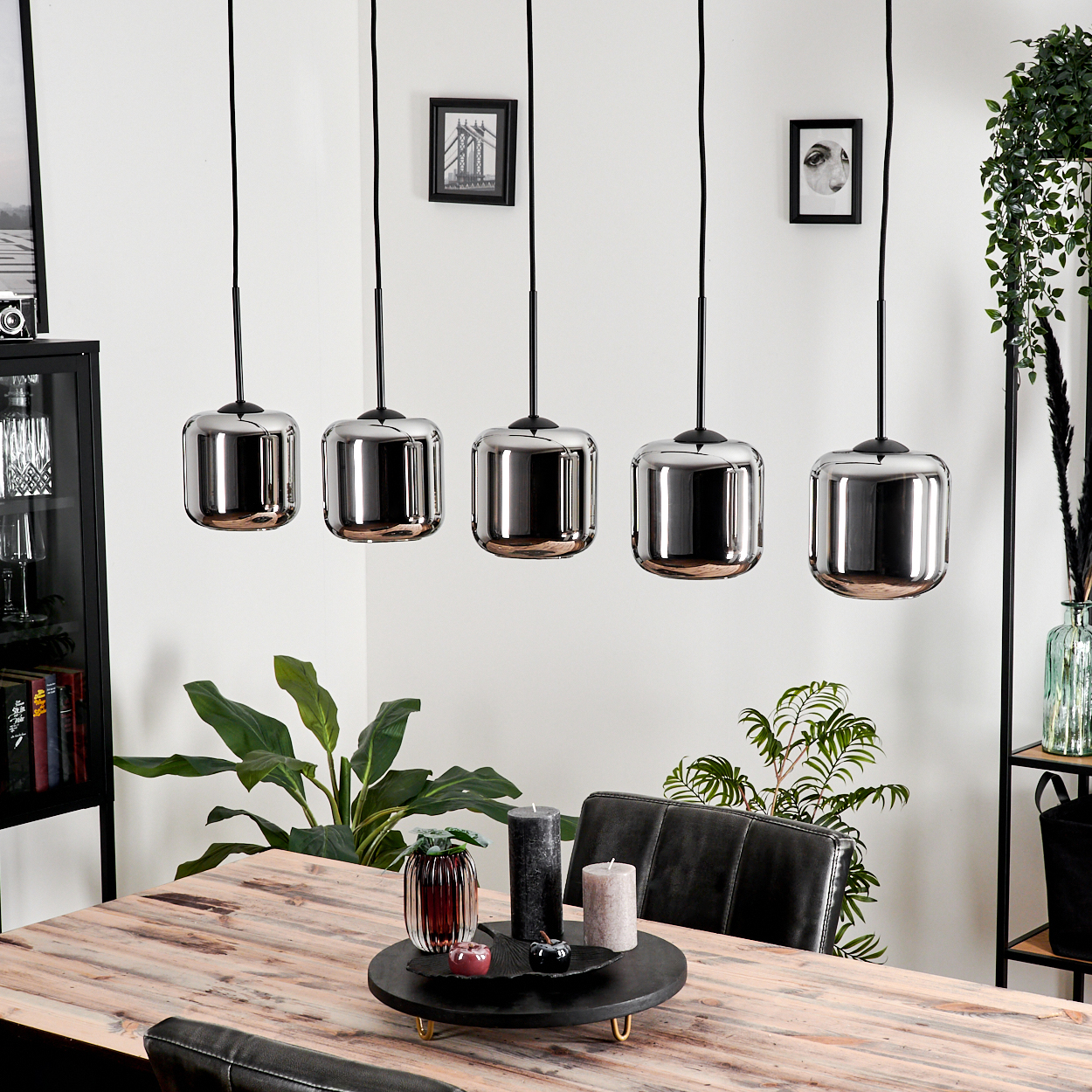 Esstisch mit fünf rechteckigen Chrom-Pendelleuchten darüber. Tisch mit Kerzen und Pflanzen gedeckt. Bilder an der Wand.