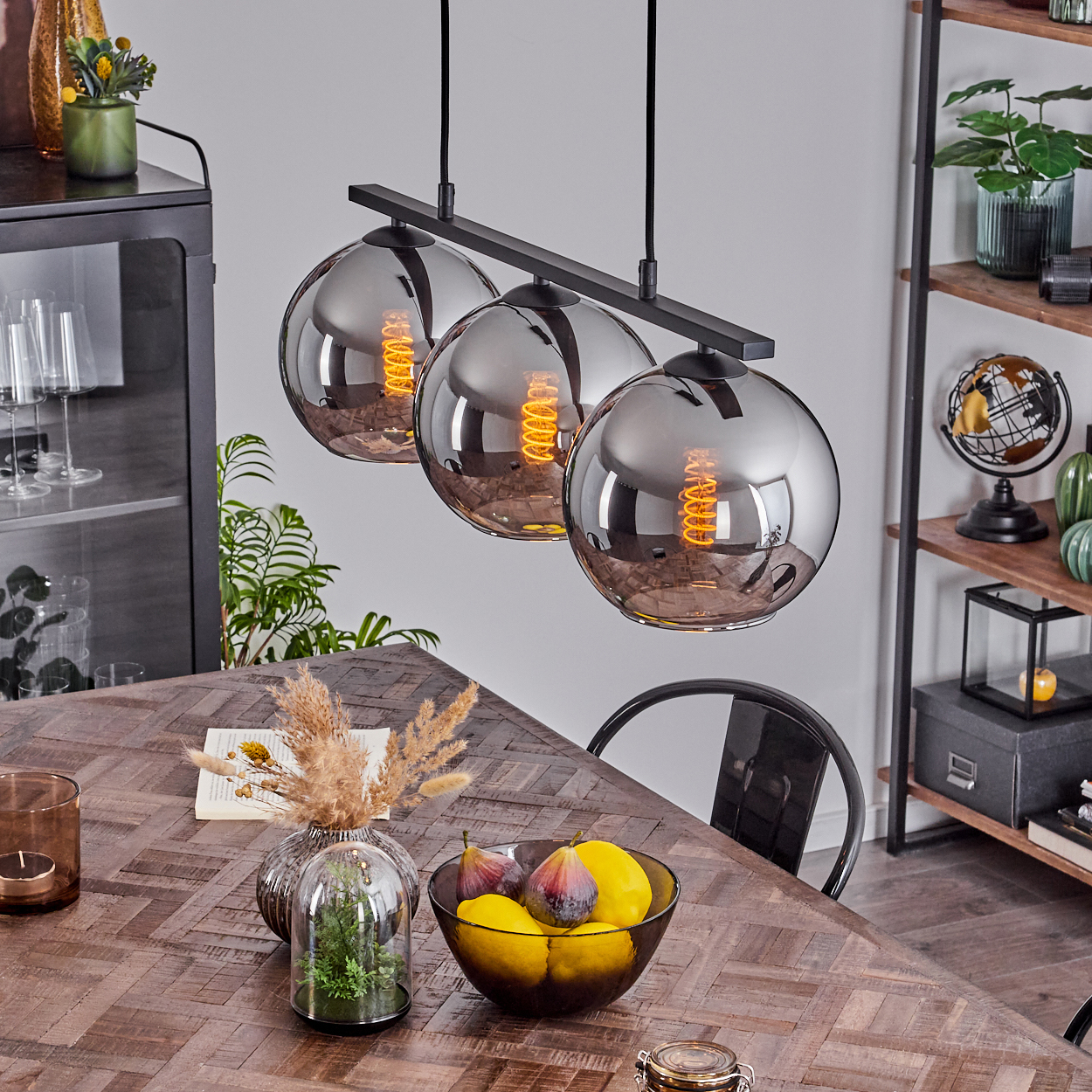 Drei Pendelleuchten mit verspiegelten Schirmen hängen über einem Holztisch. Esszimmer mit Dekoration und Obst.