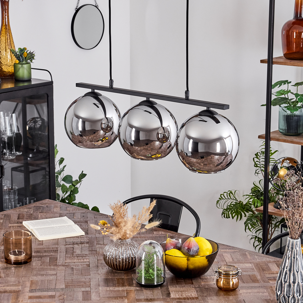 Drei Pendelleuchten mit verspiegelten Schirmen hängen über einem Holztisch. Esszimmer mit Dekoration und Obst.