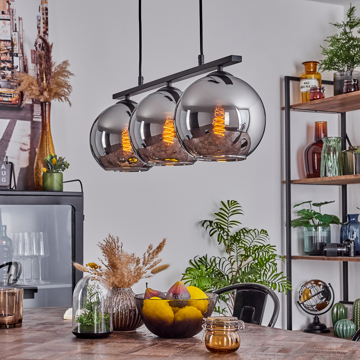 Drei silberne Kugellampen hängen über einem Tisch mit Obst. Im Hintergrund Regale und Kunstwerke.