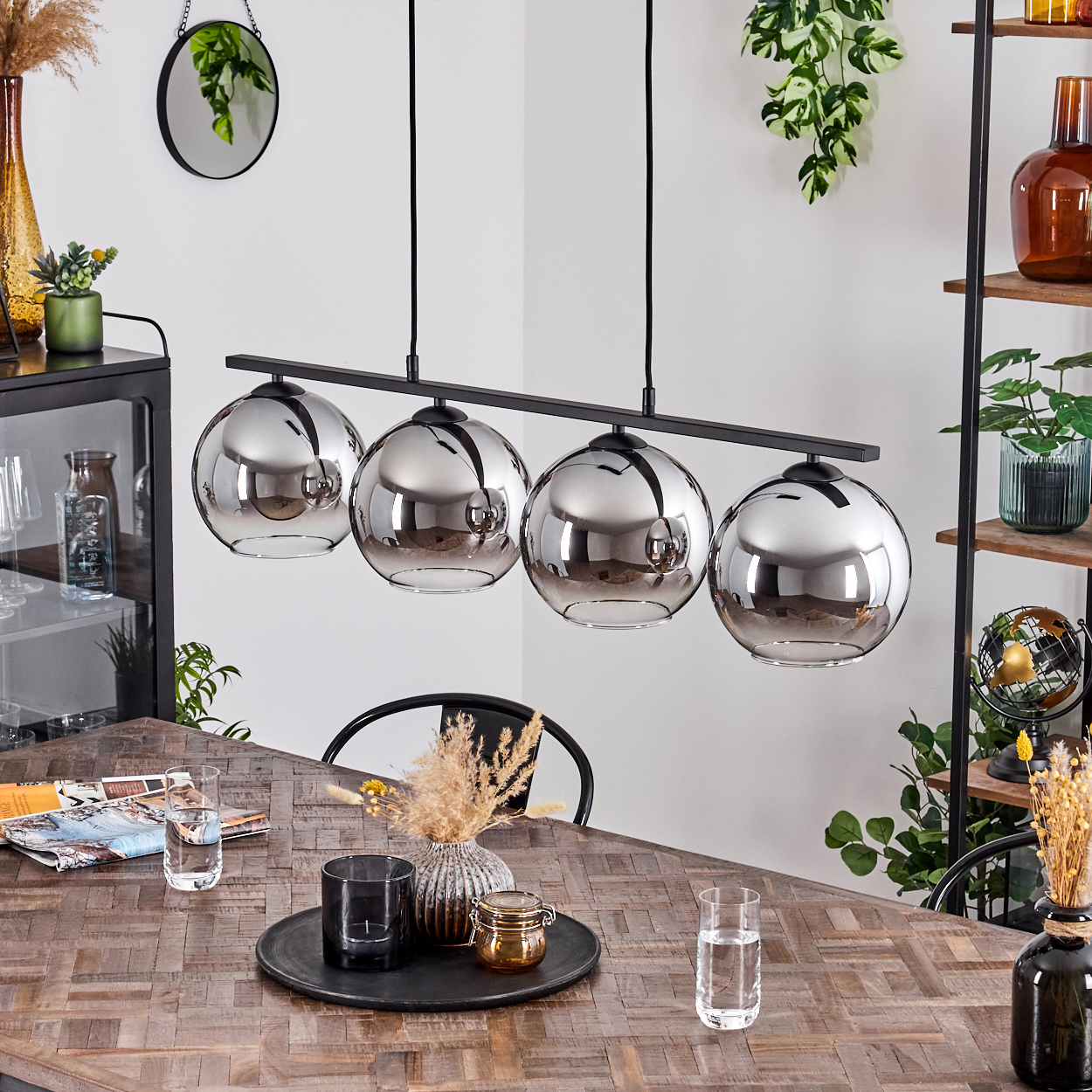 Ein Esszimmer mit Holztisch, Pendelleuchten, Pflanzen und Dekoration. Vier Glaslampen hängen über dem Tisch.