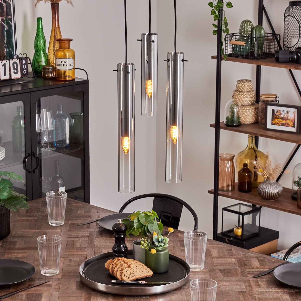 Ein Esstisch mit Gläsern, Tellern und drei Hängelampen mit Glaszylindern. Ein Regal im Hintergrund.