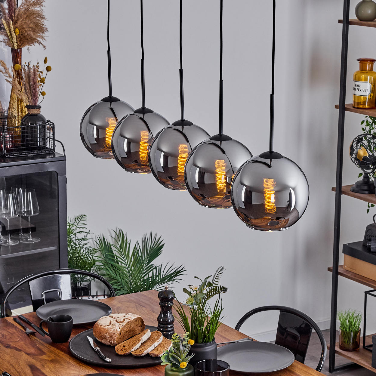 Esszimmer mit Tisch, fünf Pendelleuchten, Pflanzen und Dekorationsartikeln. Glaskugelleuchten mit Glühfäden.
