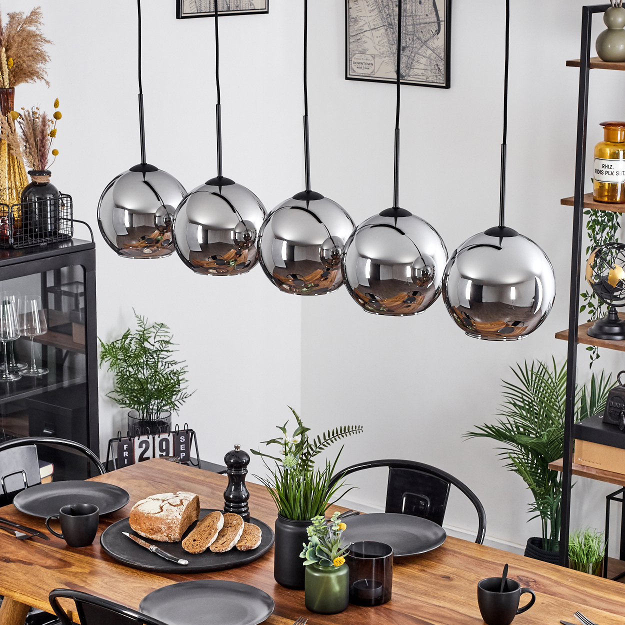 Esszimmer mit Tisch, fünf Pendelleuchten, Pflanzen und Dekorationsartikeln. Glaskugelleuchten sind sichtbar.