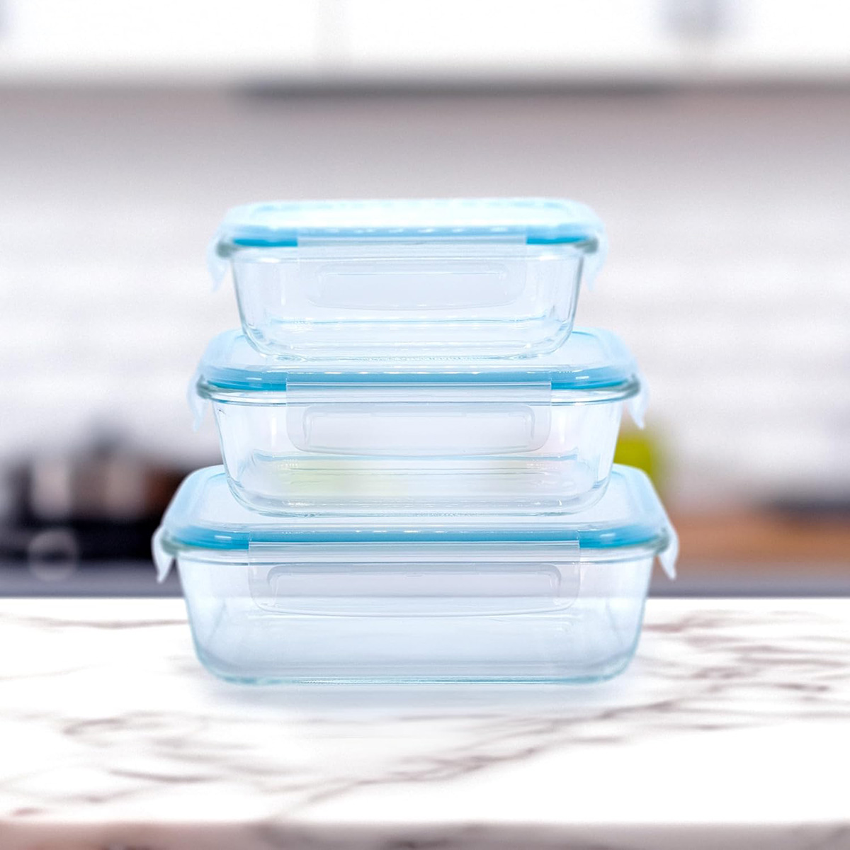 Tres recipientes de vidrio con tapas azules apilados sobre una superficie de mármol con un fondo de cocina borroso.