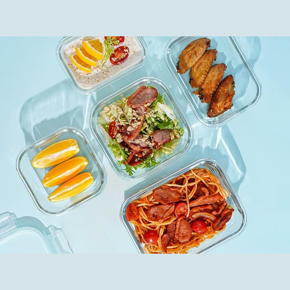 Recipientes de vidrio con diversos alimentos: ensalada, pasta, rodajas de naranja, alitas. Fondo azul.