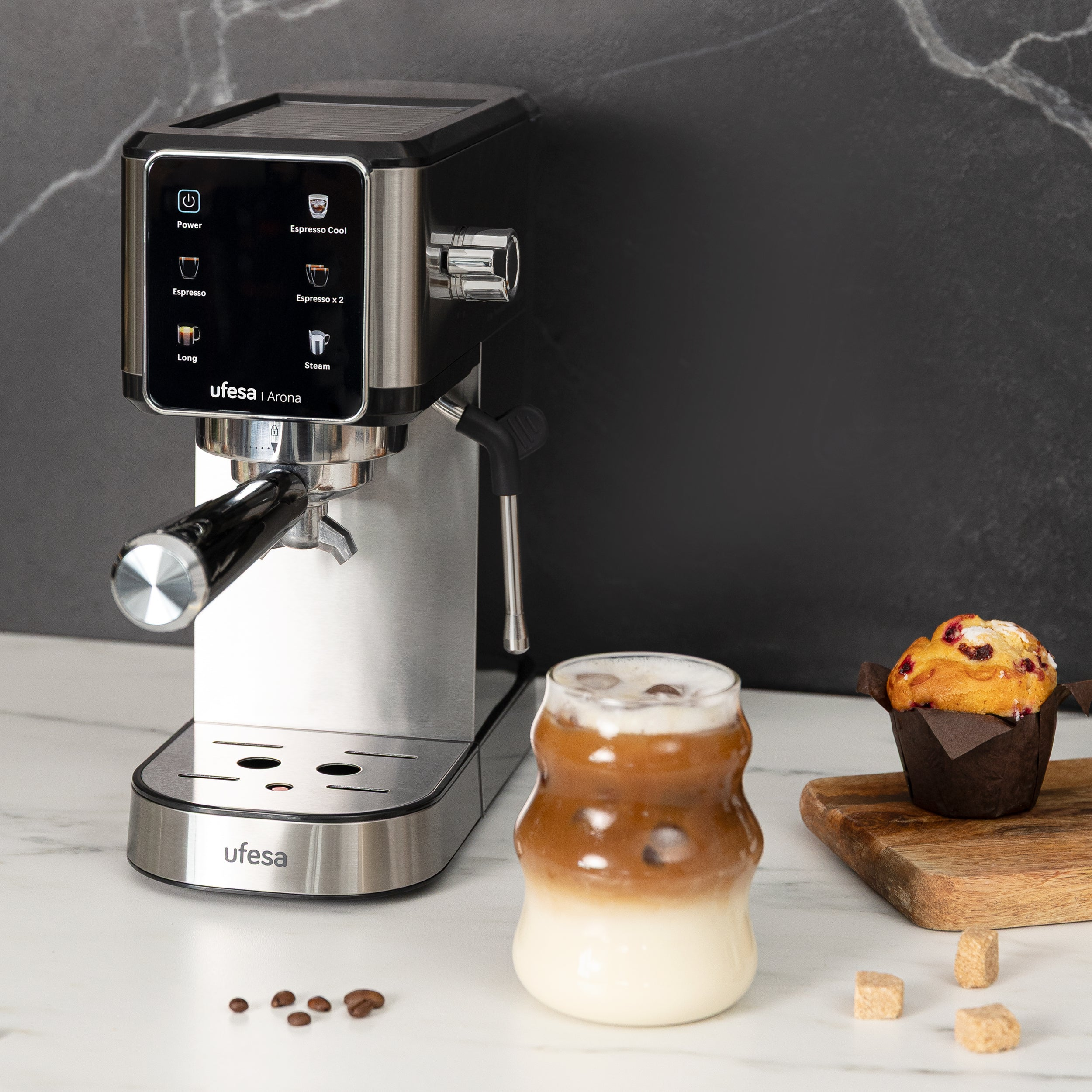 Máquina de espresso con una taza de café con leche, un muffin y granos de café en una encimera de mármol.