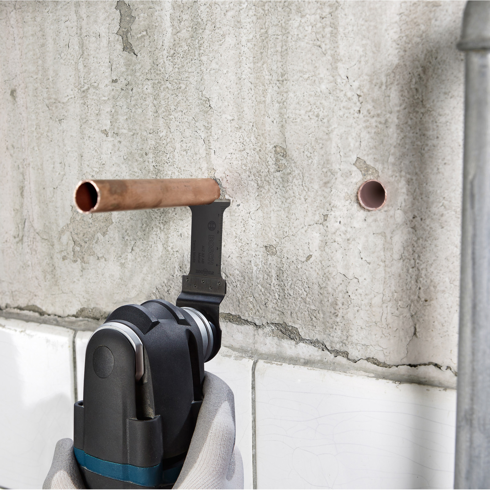 Ein Multitool-Messer schneidet durch Kupferrohre, die in einer Betonwand eingebettet sind. Ein zweites Rohr ist sichtbar.