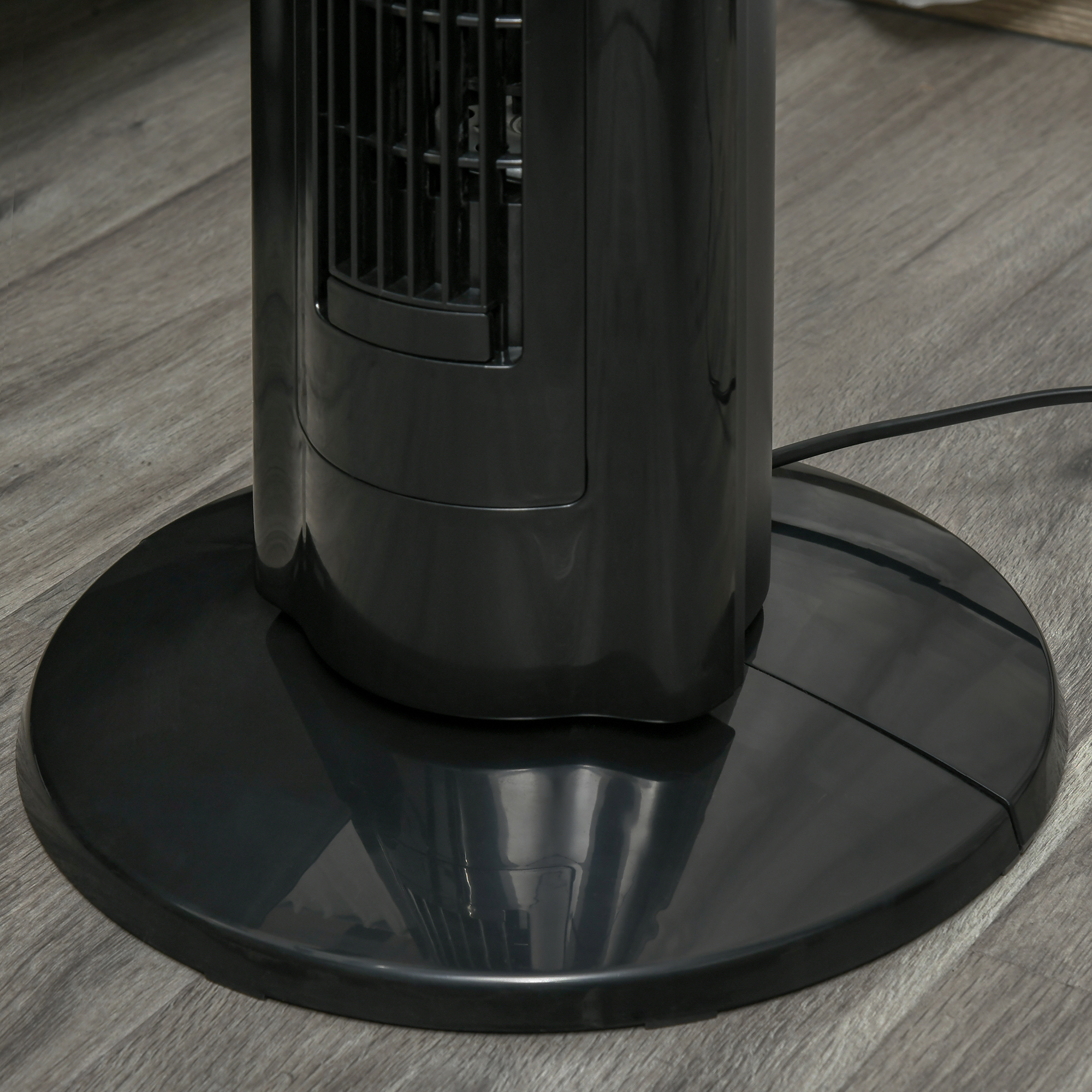 Primo piano della base rotonda e nera di un ventilatore a torre appoggiato su un pavimento effetto legno.