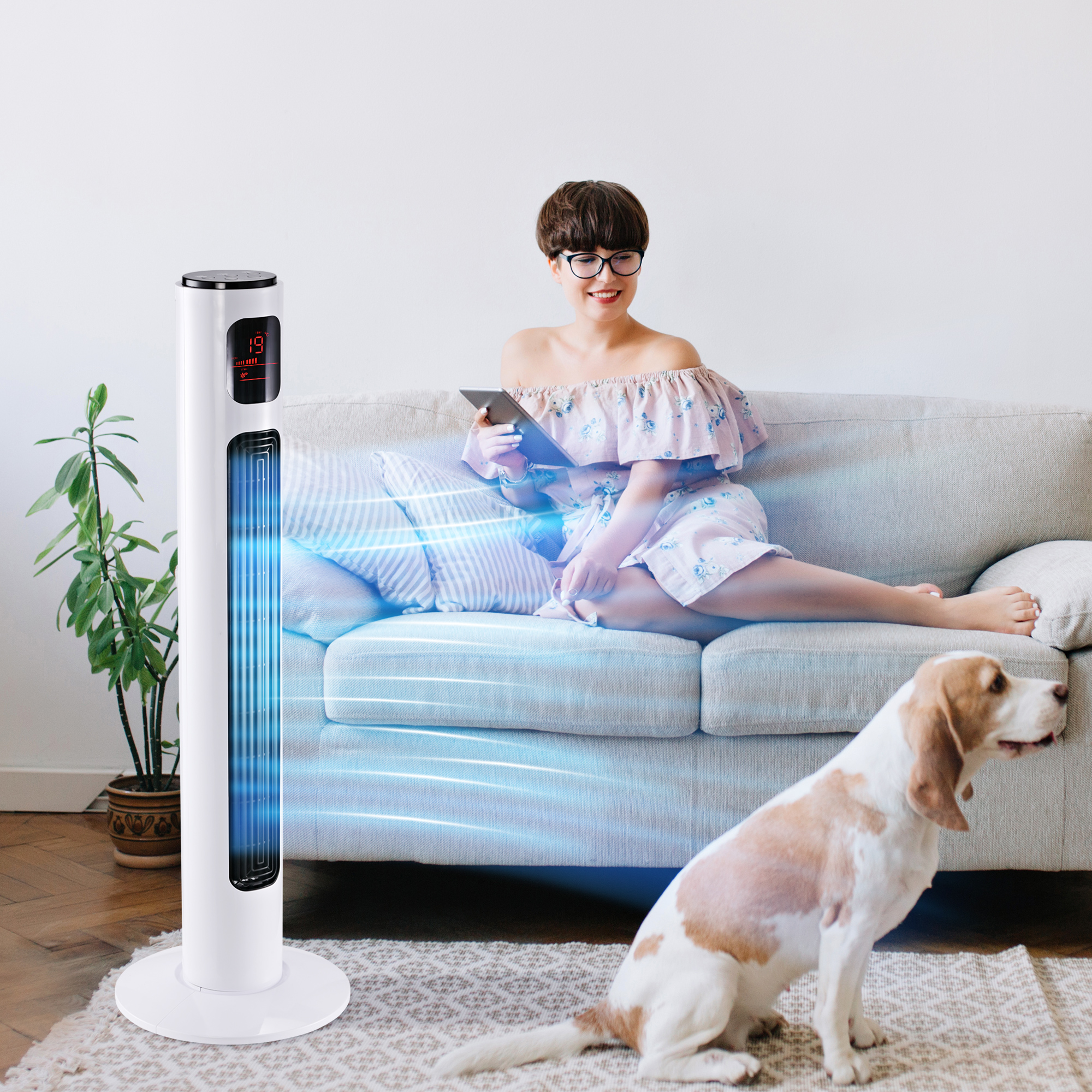 Una mujer se sienta en un sofá junto a un ventilador de torre con un perro en primer plano.