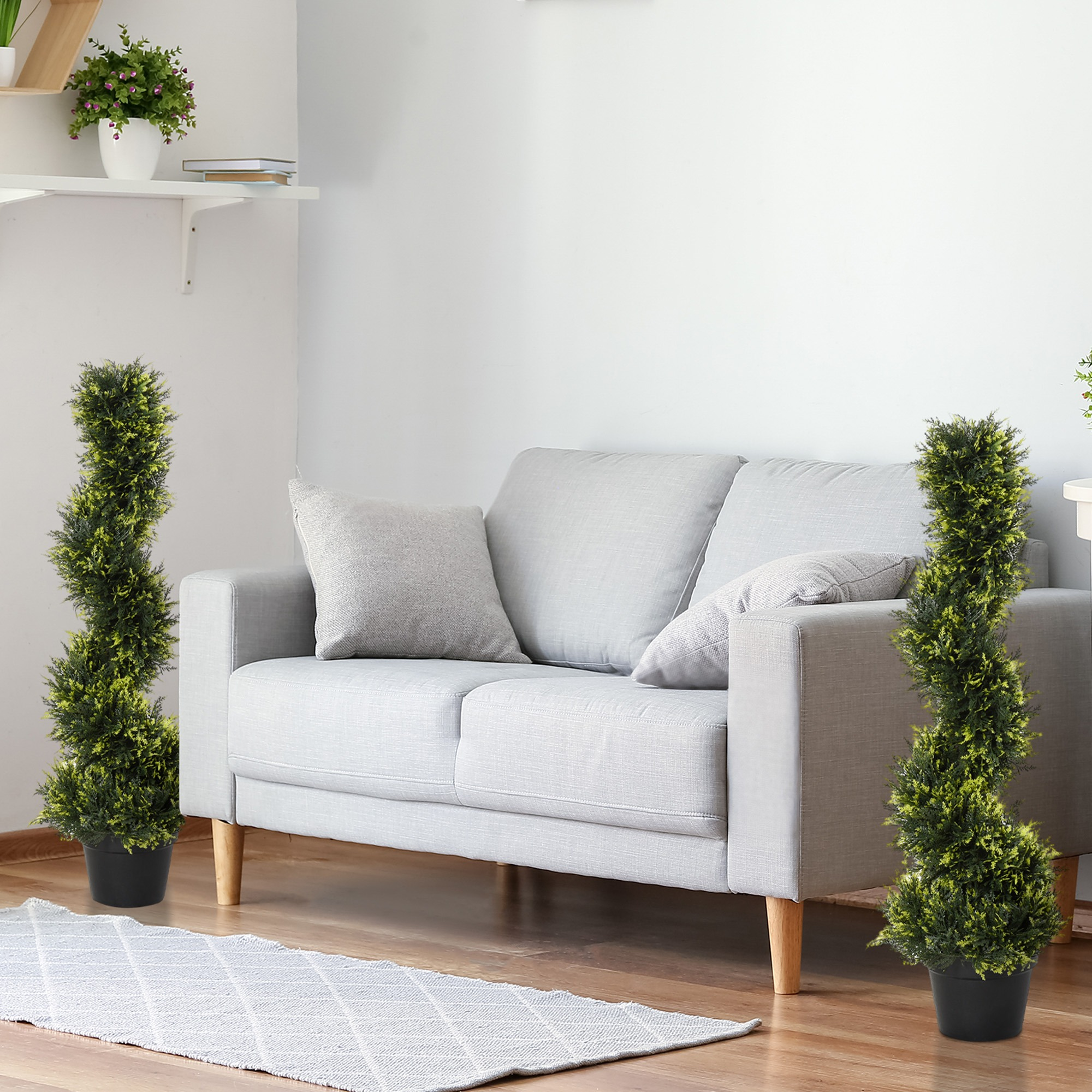 Zwei künstliche Topiaries in schwarzen Töpfen neben einem grauen Sofa auf einem Holzboden.