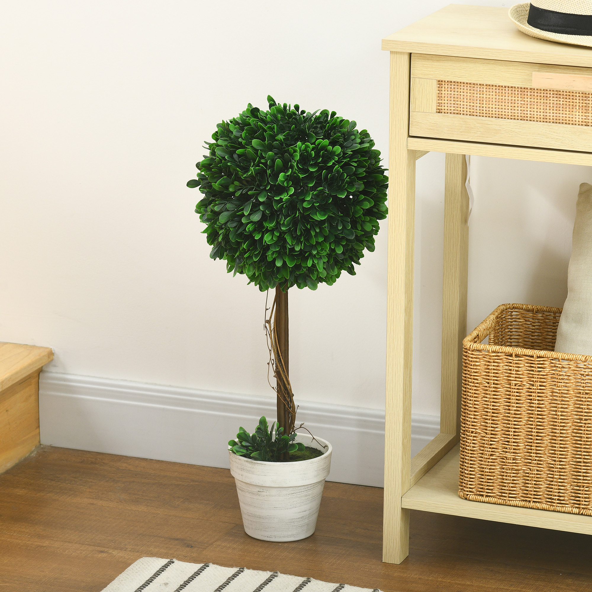 Kleiner Formschnittbaum in einem weißen Topf. Er steht neben einem Beistelltisch und einem geflochtenen Korb. Der Hintergrund ist weiß.