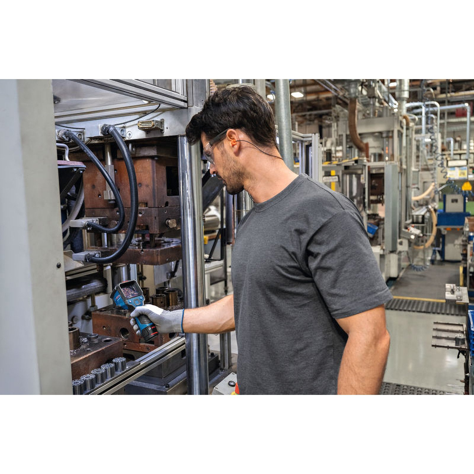 Mann in der Fabrik, mit Schutzbrille, Ohrstöpseln, hält ein Werkzeug in der Nähe von Maschinen. Industrielle Umgebung.