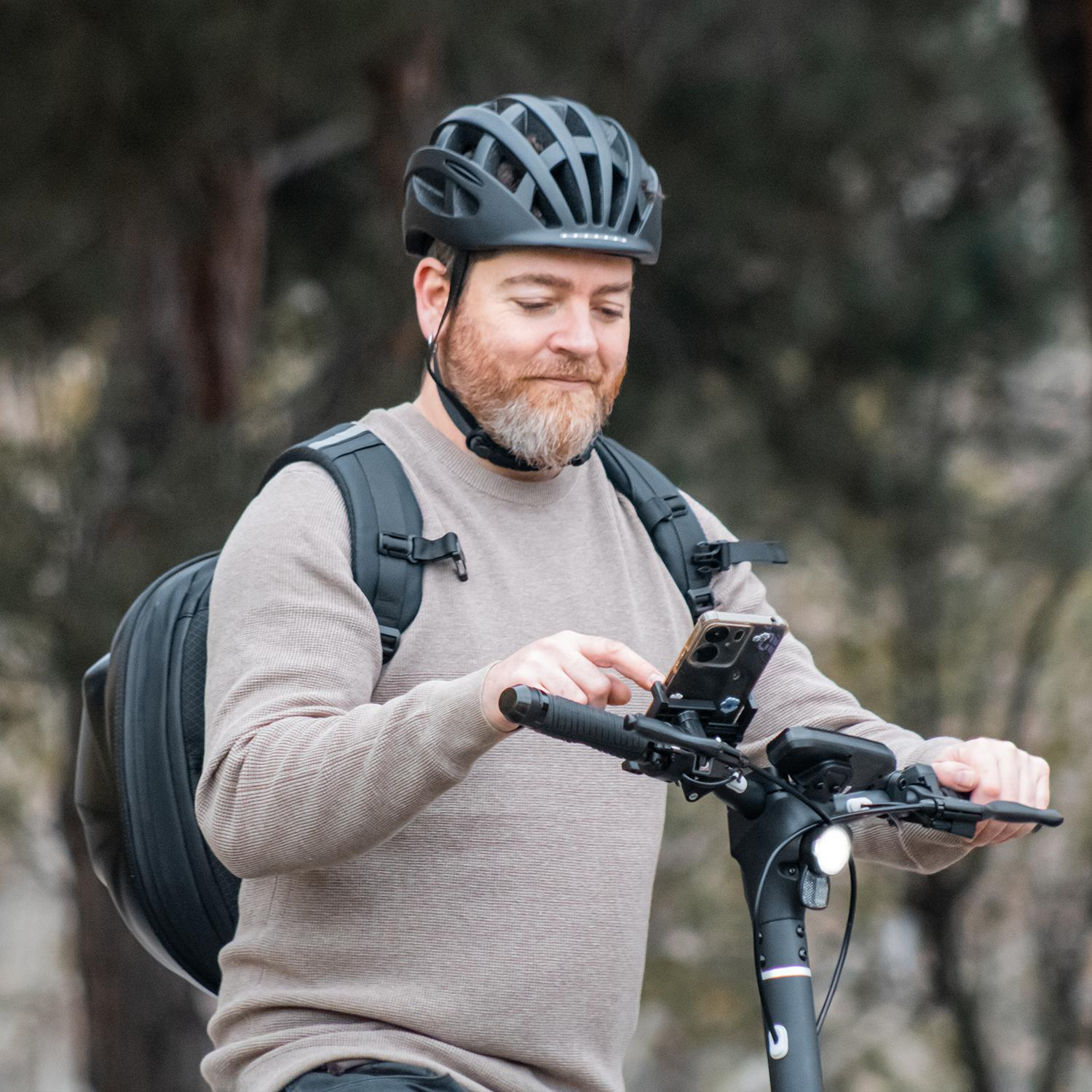 Un hombre usando un teléfono en un scooter eléctrico. Lleva casco y mochila.