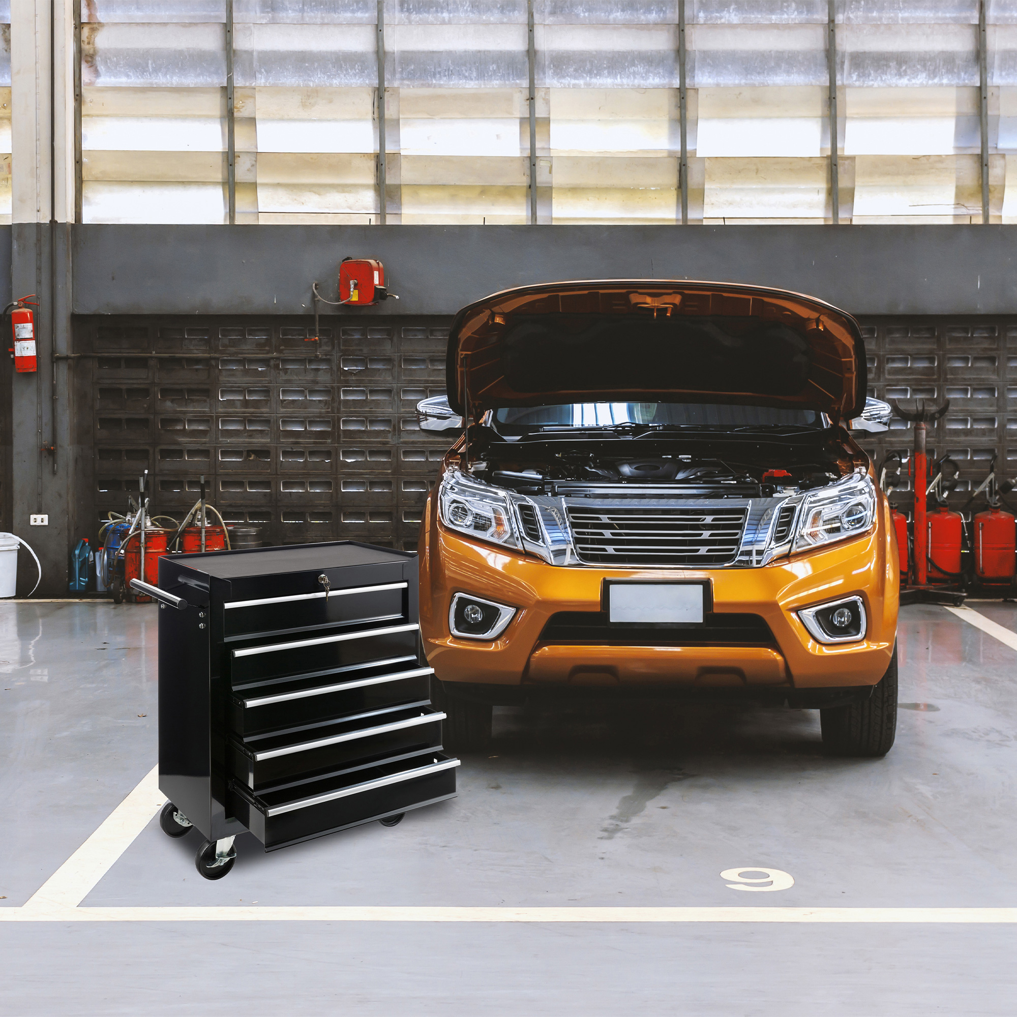 Eine schwarze Werkzeugkiste auf Rädern in einer Garage. Im Hintergrund steht ein orangefarbenes Auto mit offener Motorhaube.