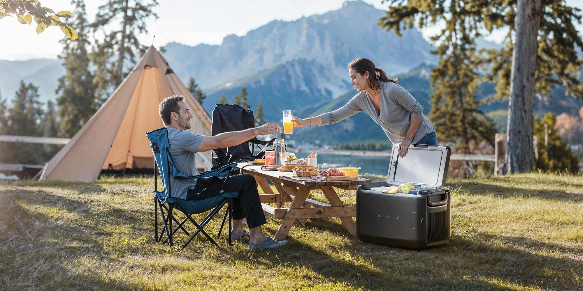 Ein Mann und eine Frau bei einem Picknick mit einer Kühlbox. Sie stoßen mit Gläsern an, im Hintergrund ein Zelt.