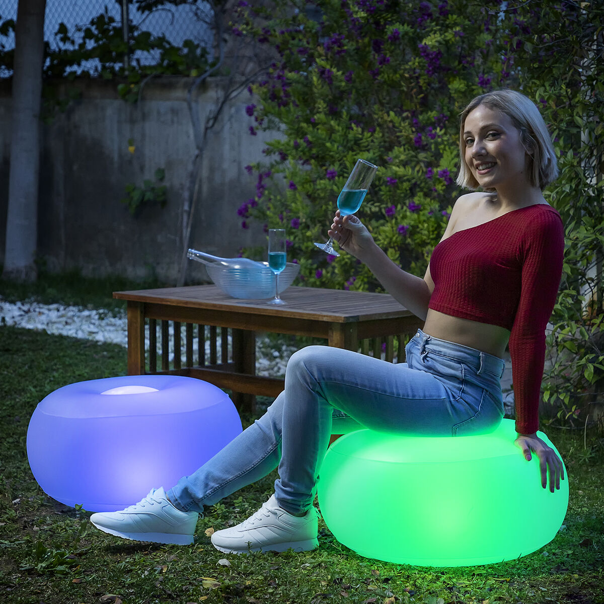 Une femme en haut rouge et jean assise sur un pouf vert lumineux, tenant un verre.