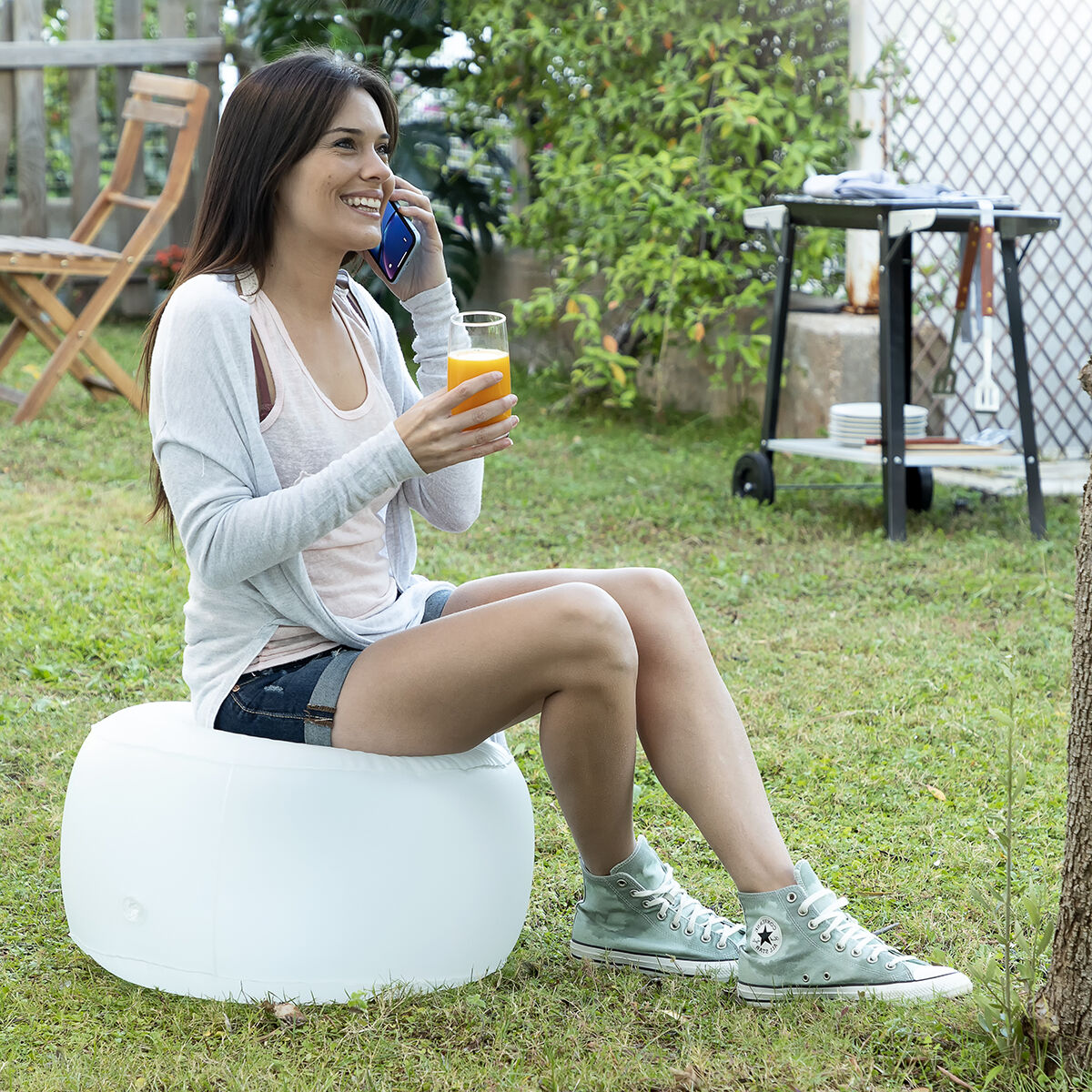 Une femme assise sur un pouf blanc dans un jardin, parle au téléphone et tient une boisson.