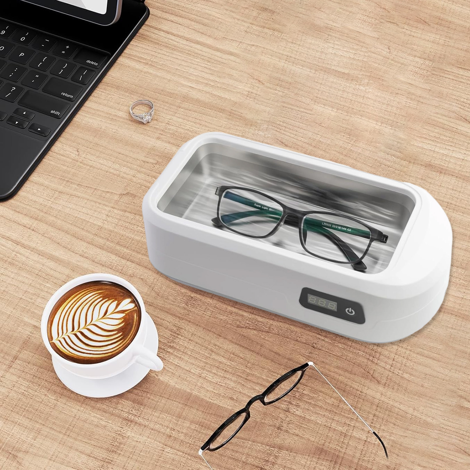 Ultraschallreiniger mit Brille, Ring und Kaffeetasse auf einem Holztisch.