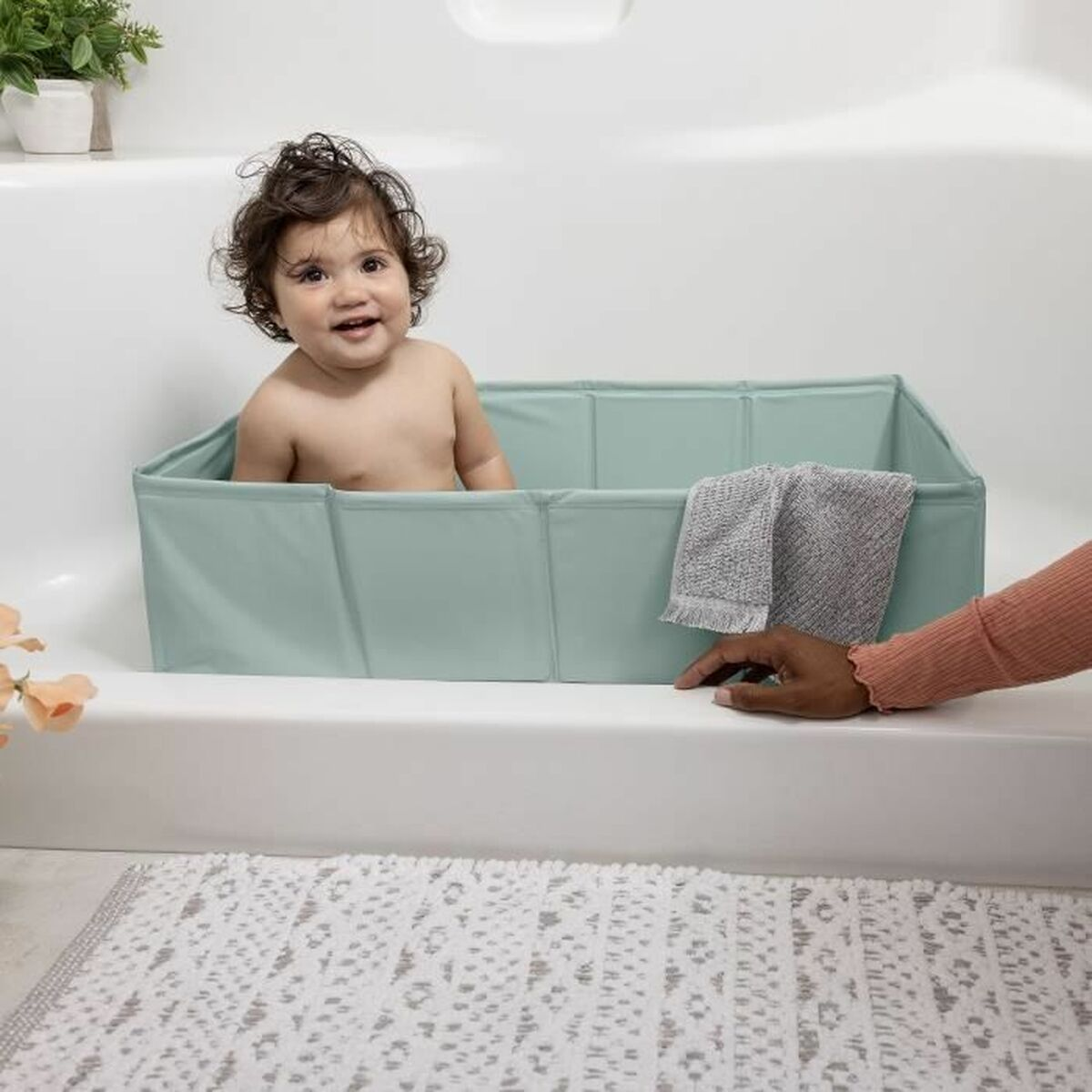 Ein Baby sitzt lächelnd in einer Babywanne in einem Badezimmer. Eine Hand greift von der Seite aus.