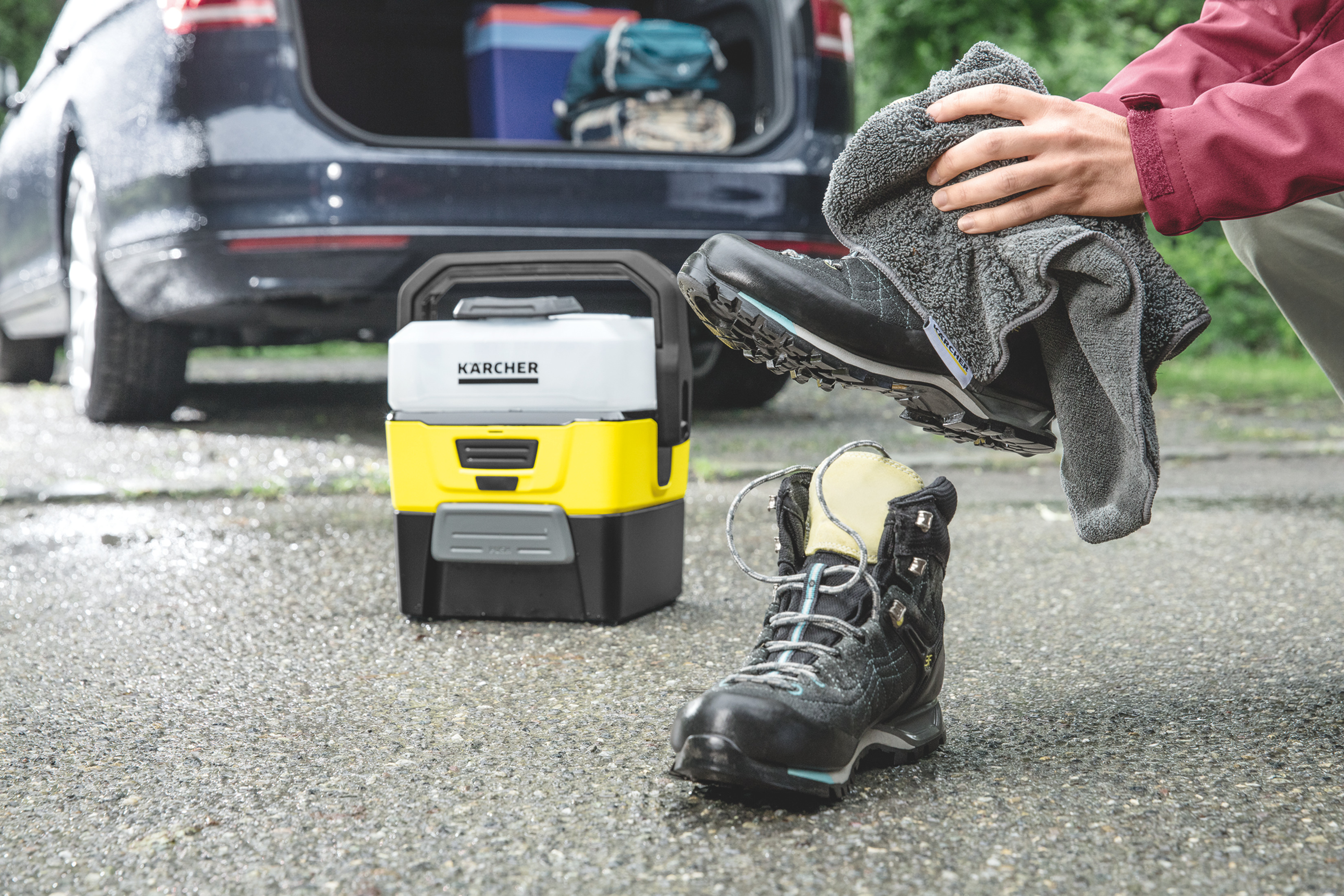 Una persona pulisce uno scarpone da trekking con un asciugamano grigio. Un pulitore portatile e un bagagliaio aperto sullo sfondo.