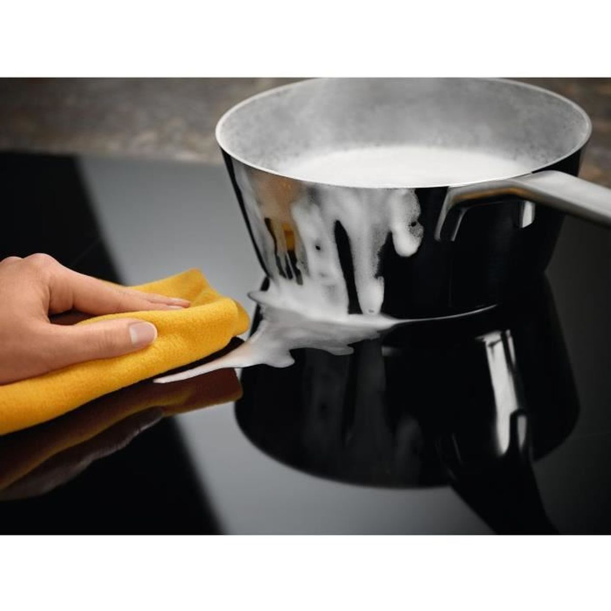 Una mano che pulisce il latte versato da un piano cottura a induzione nero con una pentola sopra.
