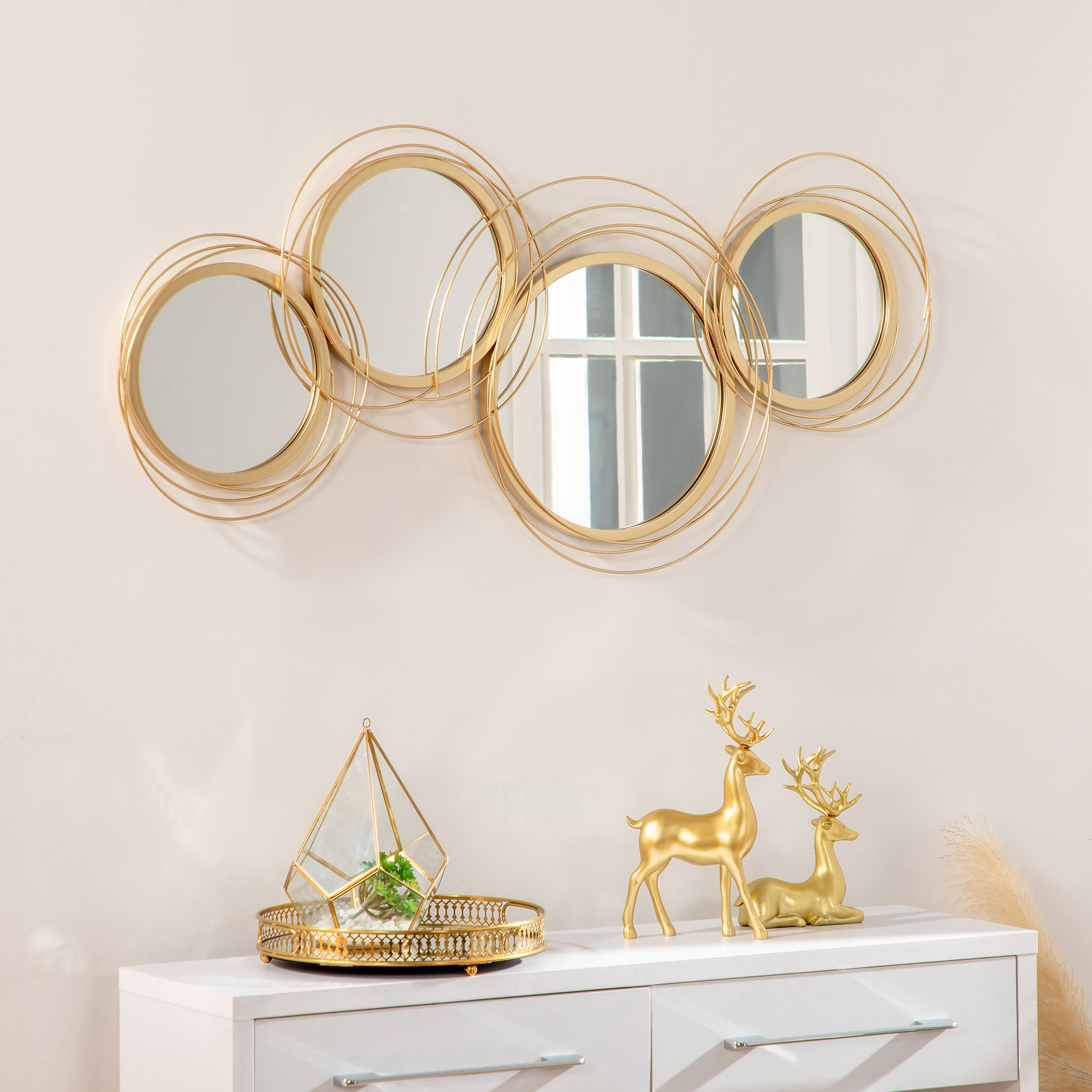 Eine weiße Kommode mit goldenen Hirschen und einem Terrarium. Darüber sind vier goldene Kreisspiegel.