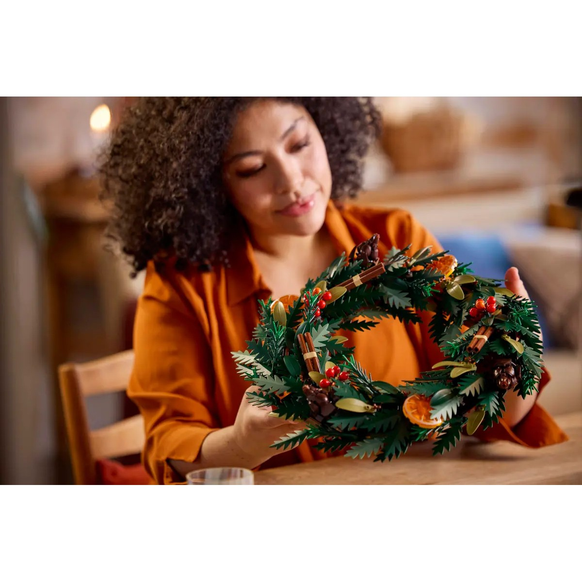 Een vrouw met krullend haar houdt een decoratieve krans vast, versierd met groene bladeren en bessen.
