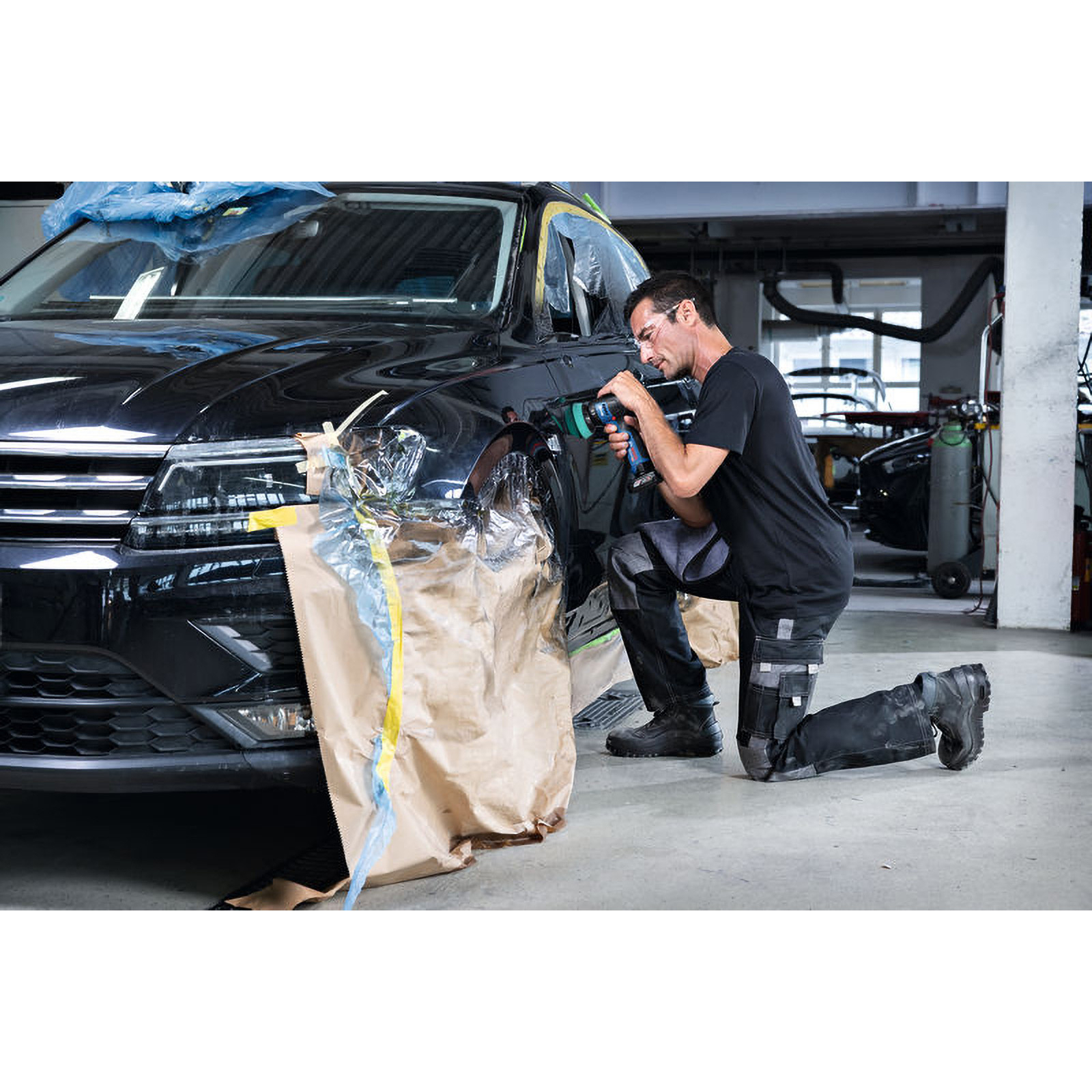 Mann kniet und poliert das Schutzblech eines schwarzen Autos. Das Auto ist abgeklebt, in einer Garage.