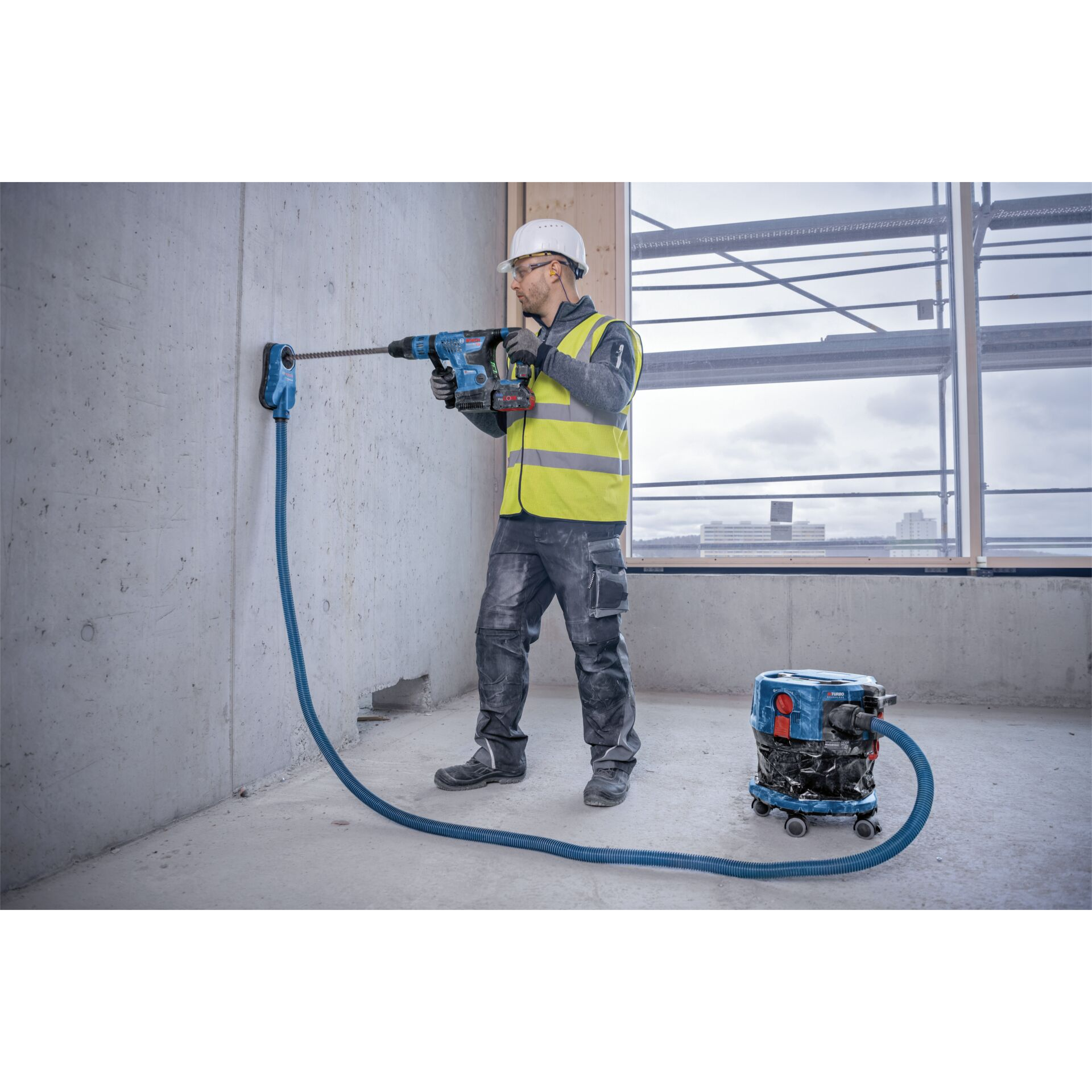 Ein Bauarbeiter bohrt mit einer Maschine eine Wand, die mit einem Staubsauger verbunden ist.