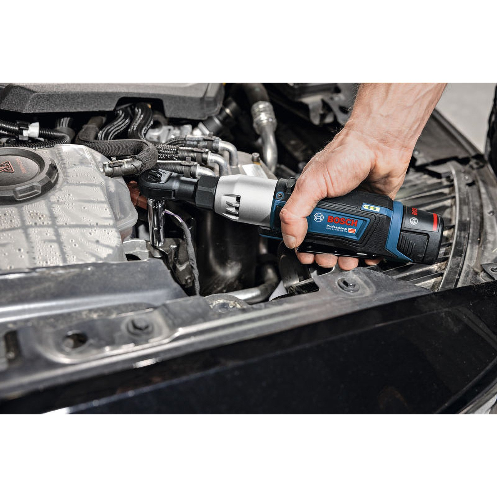 Eine Hand hält einen schwarz-blauen Bosch-Ratschenschlüssel in der Nähe eines Automotors. Der Motor ist dunkel.