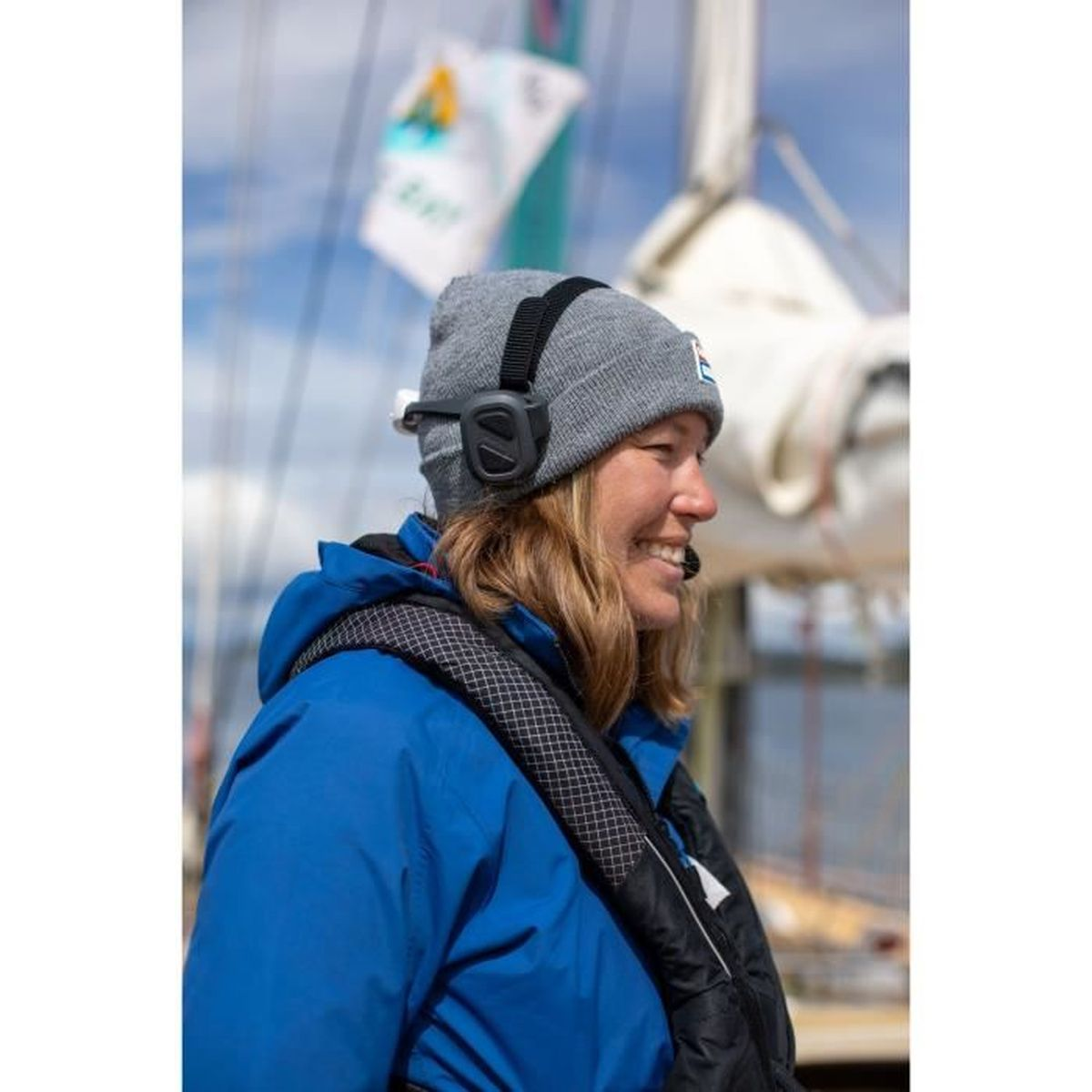 Donna su una barca con un berretto grigio e cuffie, sorridente. Sfondo: acqua e cielo.