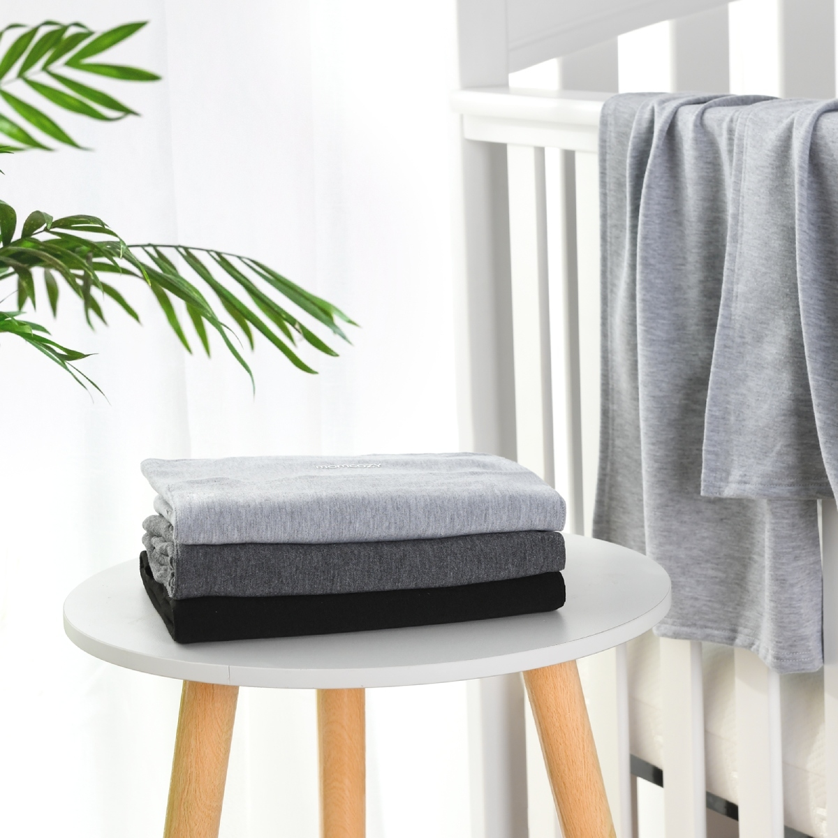 Drei gefaltete graue, dunkelgraue und schwarze Decken auf einem Tisch neben einem Kinderbett. Weißer Hintergrund.