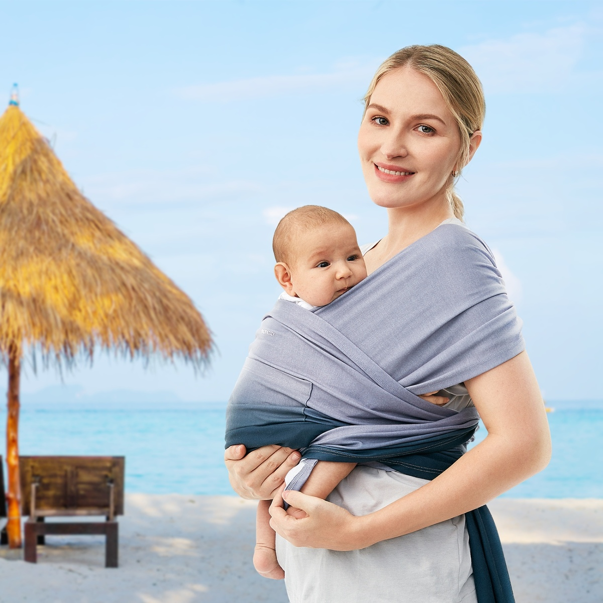 Frau hält Baby in Trage am Strand. Strandhintergrund mit Strohschirm und Meer.