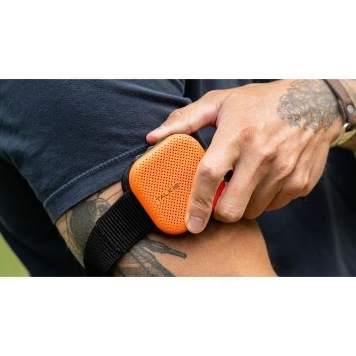 Il braccio di un uomo ha un piccolo altoparlante arancione legato. Dice 'TALKIE'. Il braccio ha un tatuaggio e un braccialetto.