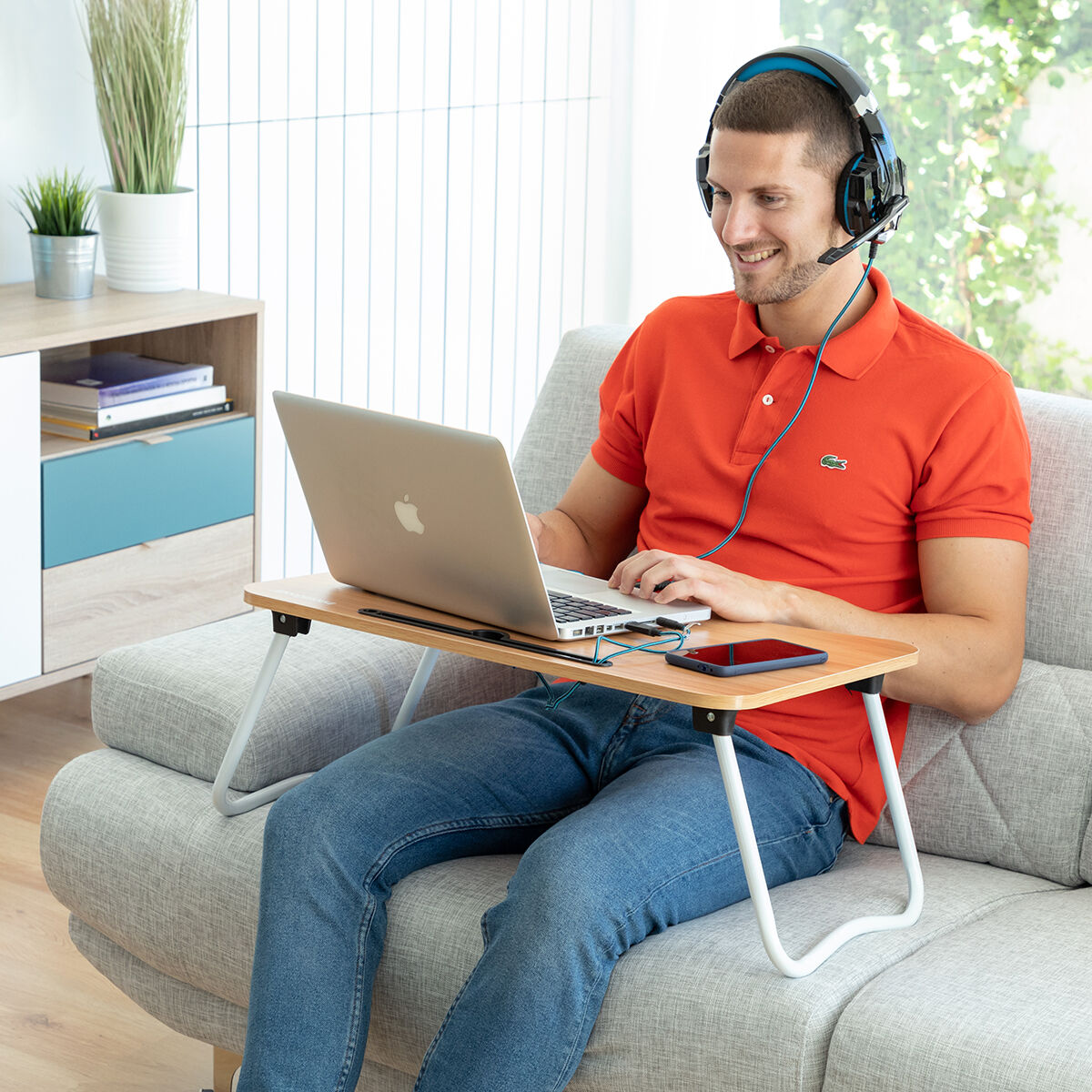 Ein Mann mit Headset, der an einem Laptop auf einem Schoßschreibtisch arbeitet und auf einem Sofa sitzt.