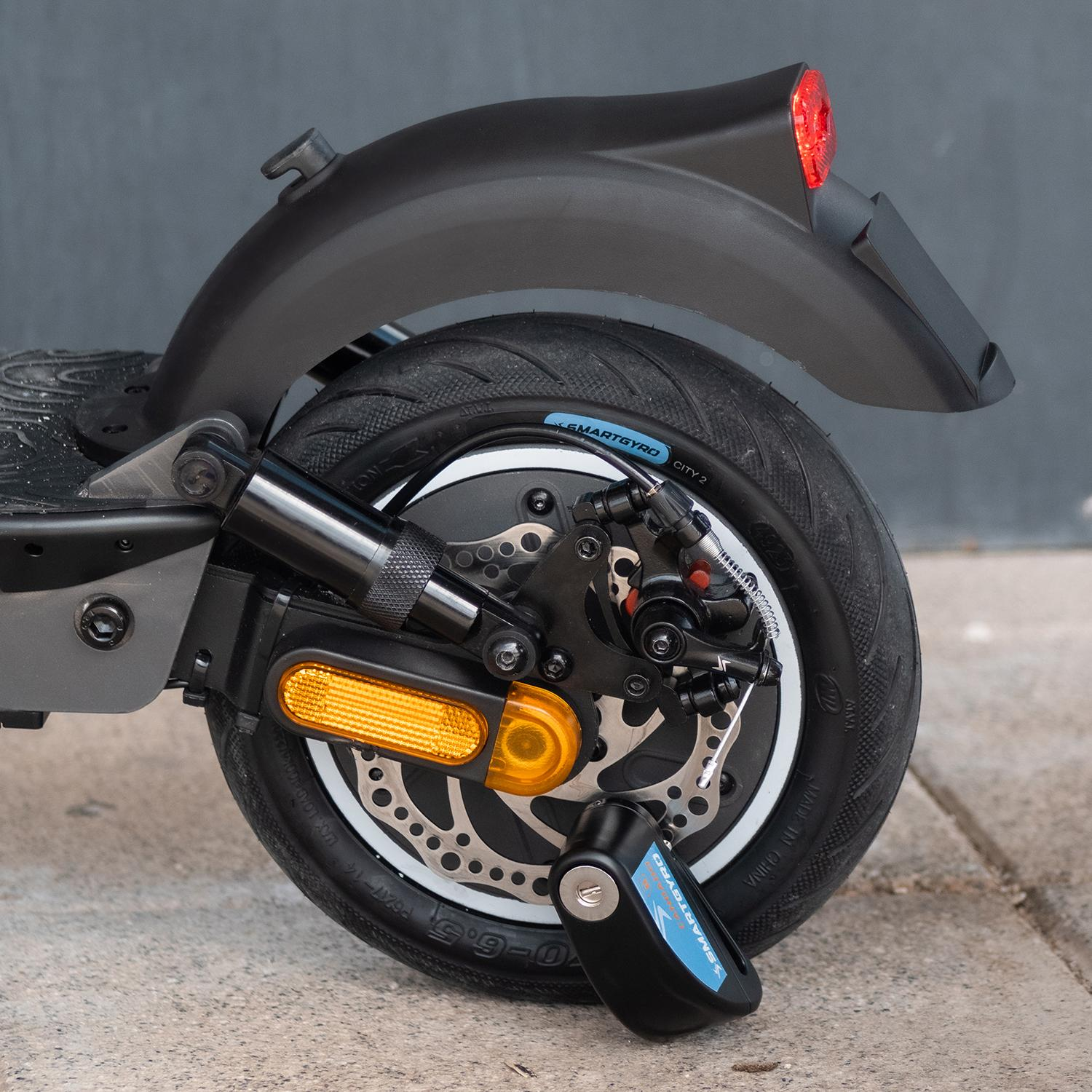 Primer plano de una rueda de scooter, con cerradura. Cuenta con neumático negro, freno de disco y un reflector lateral.