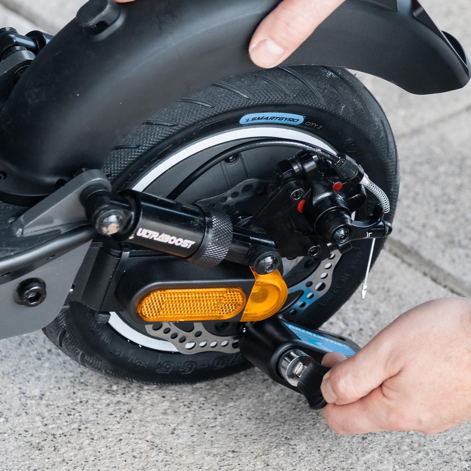 Manos instalando un candado Candado Smartgyro en una rueda de scooter. La rueda tiene freno de disco y un reflector naranja.