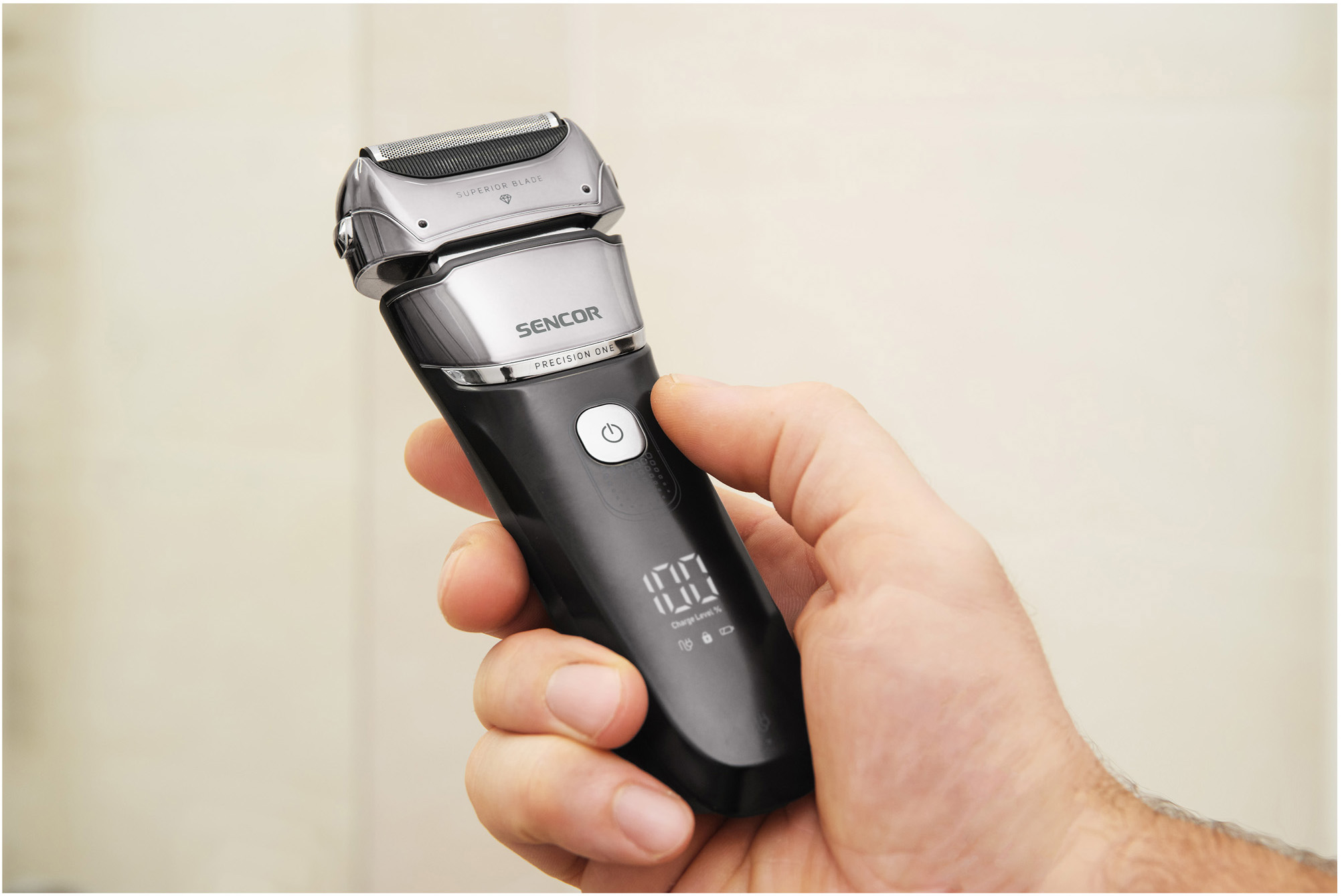 Una mano tiene in mano un rasoio elettrico Sencor nero e argento con un display digitale che mostra "100".