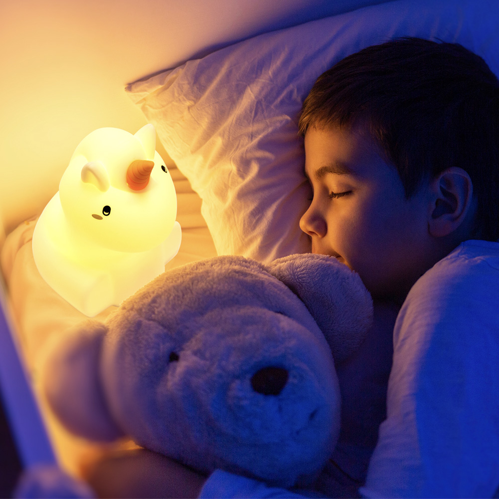 Un ragazzo dorme con una lampada a forma di unicorno e un orsacchiotto sul letto.