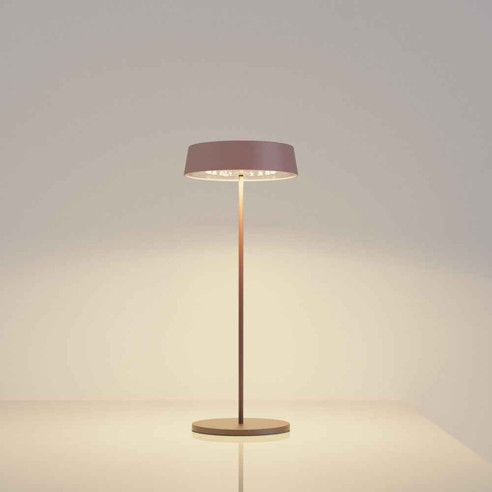 Una lampada da tavolo con paralume malva e supporto color rame. La luce è accesa.
