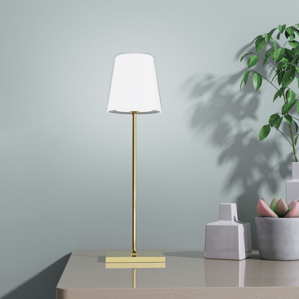 Una lampada dorata su un tavolo. Un paralume bianco in cima. Pianta e vaso sullo sfondo.