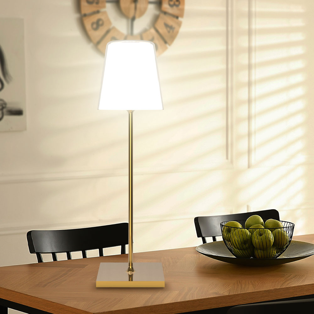 Una lampada con base dorata e paralume bianco su un tavolo di legno. Orologio e ciotola con mele visibili.