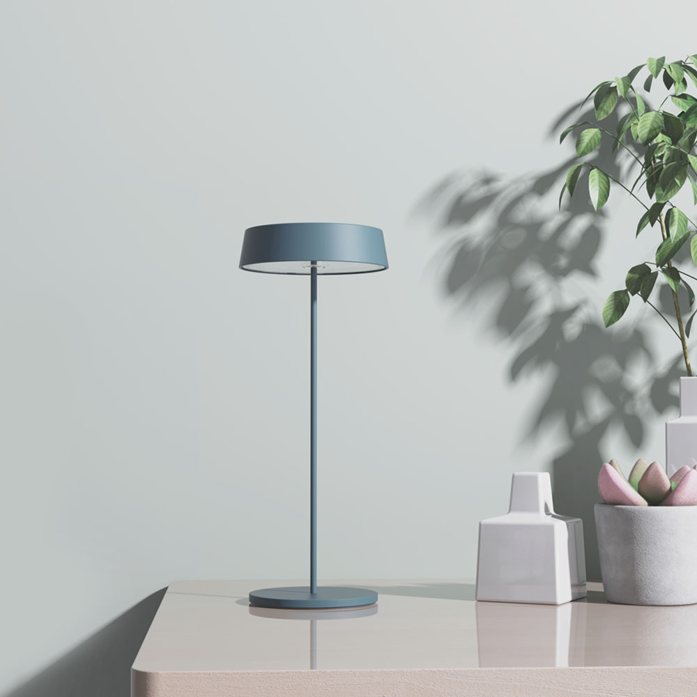 Una lampada con paralume e stelo blu, su un tavolo con un vaso bianco e una pianta con foglie verdi.