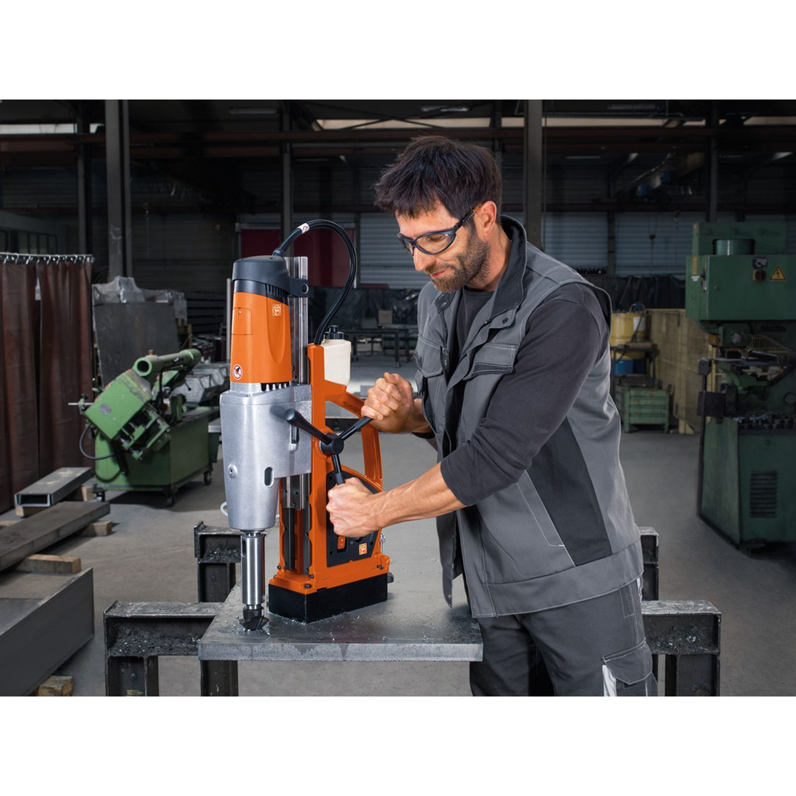 Ein Mann mit Schutzbrille bedient eine orangefarbene und silberne Bohrmaschine auf einer Metalloberfläche.
